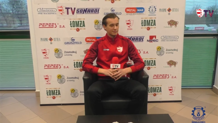 Piłkarz w dresach klubu ŁKS Łomża siedzi na fotelu w budynku klubowym z mikrofonem w rękach. W tle jest ścianka z logami sponsorów klubu ŁKS Łomża.