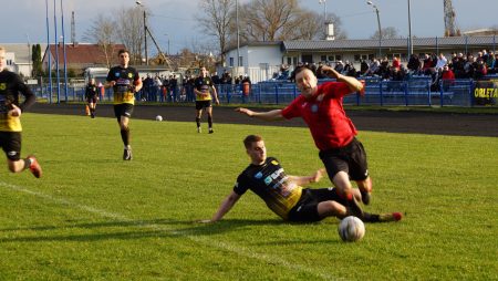 Piłkarz podczas meczu piłkarskiego wślizgiem próbuje odebrać piłkę zawodnikowi Pogoni Łapy. Ten upada na boisko. Na trybunach kibice.