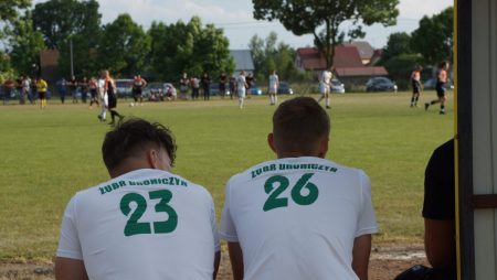 Na pierwszym planie dwóch piłkarzy w białych koszulkach Żubra Drohiczyn widzianych tyłem. Na drugim planie mecz piłkarski.
