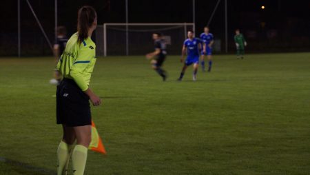 Mecz piłki nożnej, na pierwszym planie pani sędzia z chorągiewką, w tle boisko piłkarskie i walka o piłkę dwóch piłkarzy.