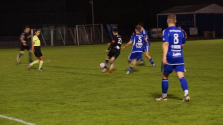 Piłkarze w czasie meczu walczą o piłkę. Grają dwie drużyny Sky Białystok i GKS Rutki. Jedni w niebieskich koszulkach, drudzy w czarnych. Sędzia patrzy na akcję.